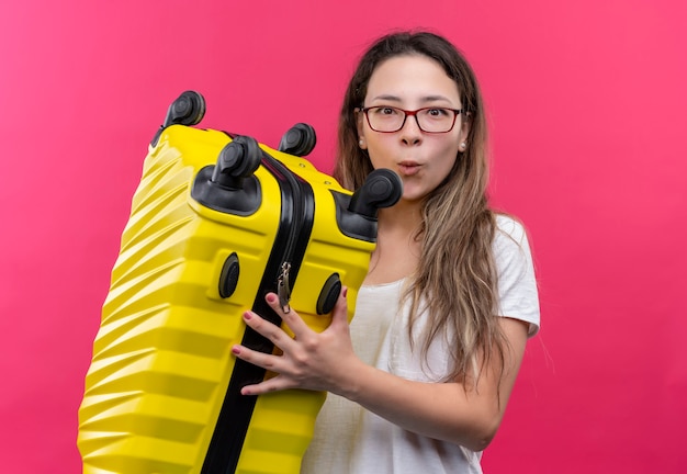 Jonge reizigersvrouw die in witte de reiskoffer van de t-shirtholding verbaasd en verbaasd over roze muur kijken