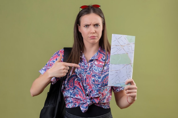 Jonge reizigersvrouw die met rode zonnebril op hoofd en met de kaart van de rugzakholding met wijsvinger aan het met fronsend gezicht over groene muur richten