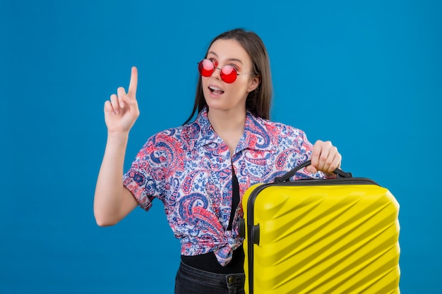 Jonge reizigersvrouw die rode zonnebril draagt die gele koffer houdt die met omhoog vinger omhoog staat kijkt zelfverzekerd met geweldig idee over blauwe achtergrond