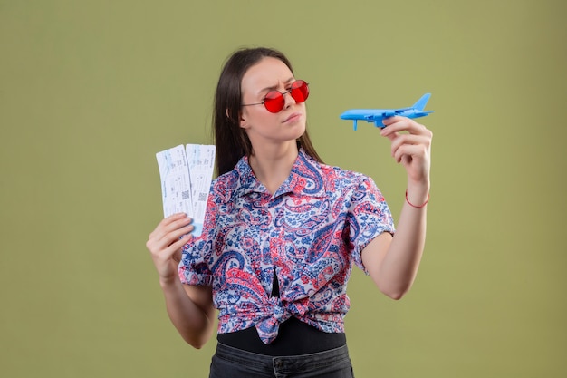 Gratis foto jonge reizigersvrouw die rode zonnebril dragen die kaartjes en stuk speelgoed vliegtuig houden die het met peinzende uitdrukking met fronsend gezicht over groene muur bekijken