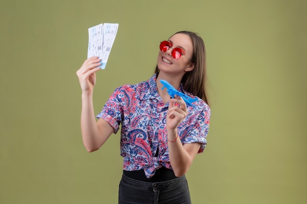 Jonge reizigersvrouw die rode zonnebril dragen die kaartjes en stuk speelgoed vliegtuig houden die opzij met gelukkig gezicht kijken die vrolijk over groene muur glimlachen