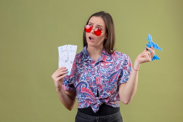 Jonge reizigersvrouw die rode zonnebril dragen die kaartjes en stuk speelgoed vliegtuig houden die opzij met peinzende uitdrukking kijken die zich over groene achtergrond bevindt