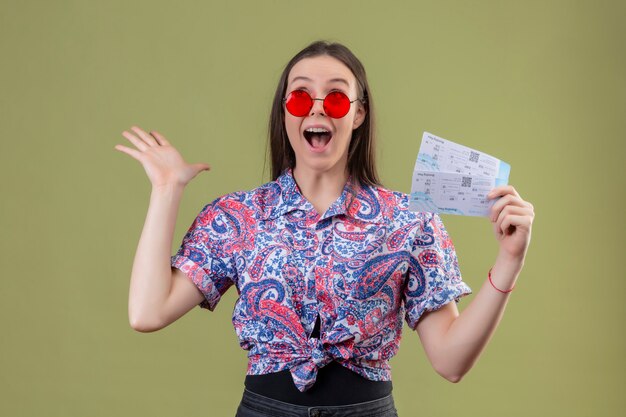 Jonge reizigersvrouw die rode zonnebril dragen die kaartjes houden die met wapens worden verbaasd en verrast over groene muur worden opgeheven