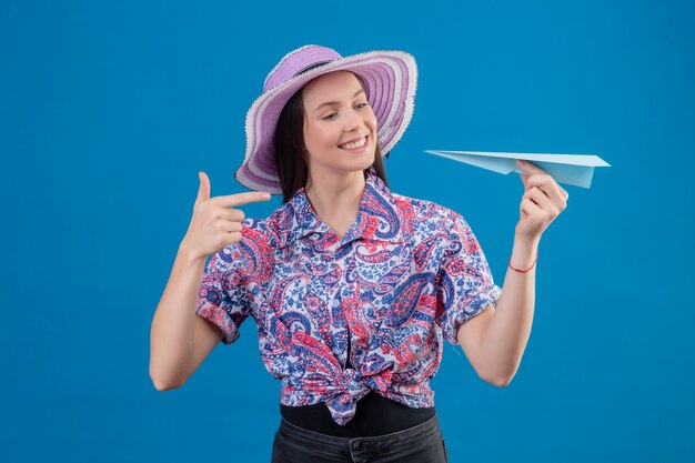 Jonge reizigersvrouw in de holdingsdocument vliegtuig van de zomerhoed die met vinger ernaar richten die met gelukkig gezicht glimlachen die zich over blauwe achtergrond bevinden