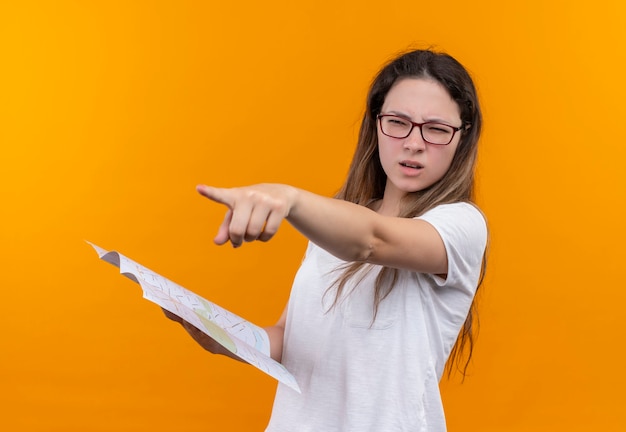 Jonge reizigersvrouw in de witte kaart die van de t-shirtholding de goede richting met vinger richt die zich over oranje muur bevindt