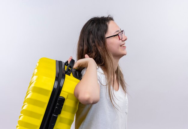 Jonge reizigersvrouw in de witte koffer die van de t-shirtholding zijwaarts glimlachend positief en gelukkig over witte muur staat