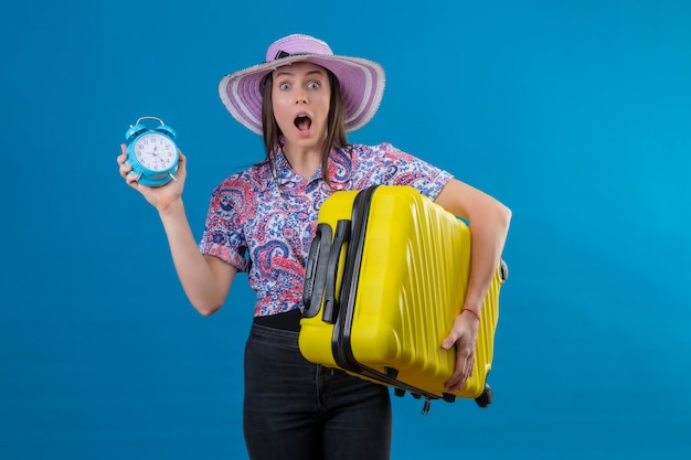 Gratis foto jonge reizigersvrouw in de zomerhoed met de gele wekker van de kofferholding die met schaamte voor fout wordt geschokt, uitdrukking van vrees over blauwe muur