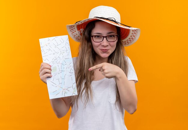 Jonge reizigersvrouw in wit t-shirt die de holdingskaart van de zomerhoed dragen die met vinger ernaar richt die met gelukkig gezicht glimlacht dat zich over oranje muur bevindt