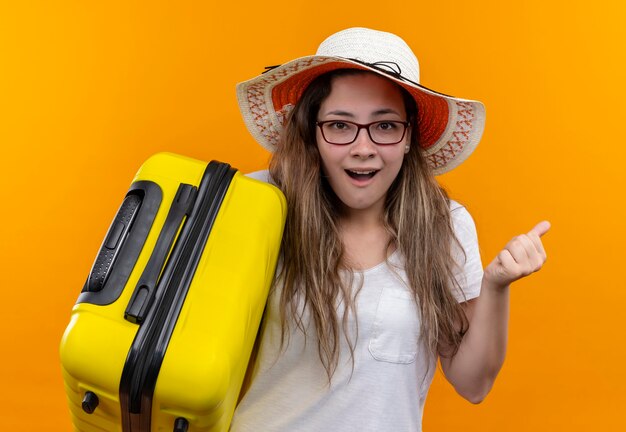 Jonge reizigersvrouw in wit t-shirt die de koffer van de de holdingskoffer van de zomerhoed dragen opgewonden en gelukkige gebalde vuist die zich over oranje muur bevindt