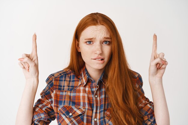 Jonge roodharige vrouw die lip bijt met een gecompliceerde blik, vingers omhoog wijst en bezorgd naar voren staart, staande over een witte muur