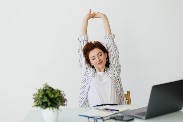 Gratis foto jonge roodharige vrouw die met haar laptop werkt en haar armen in ontspannen positie strekt