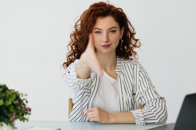 Jonge roodharige vrouw die werkt met haar laptop die haar hand uitstrekt naar de camera in begroetingsgebaar