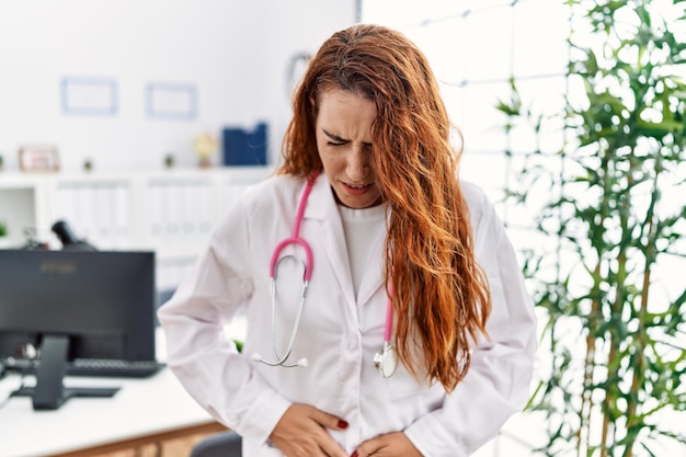Jonge roodharige vrouw in doktersuniform en stethoscoop in de kliniek met de hand op de buik omdat indigestie pijnlijke ziekte onwel pijn concept