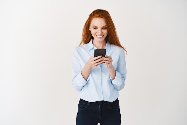 Jonge roodharige vrouw leest bericht op mobiele telefoon en glimlacht