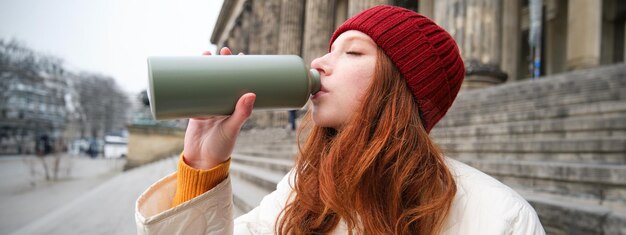 Jonge roodharige vrouwelijke toerist rust tijdens haar reis, opent thermoskan en drinkt hete thee tijdens een pauze