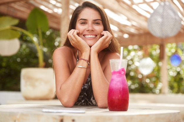 Jonge schattige vrouw met brede glimlach rust op bar tijdens zomerdag, drinkt exotisch fruit shake, geniet van goede rust in hete exotische land. Vrij lachende vrouw recreëren in stoeprestaurant.