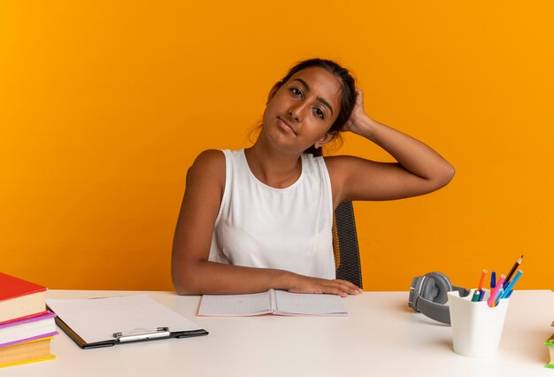 jonge schoolmeisje zit aan bureau met school tools hand op het hoofd te zetten