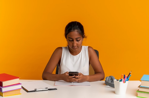 Jonge schoolmeisjeszitting aan bureau met het nummer van de schoolhulpmiddelen op telefoon die op oranje muur wordt geïsoleerd