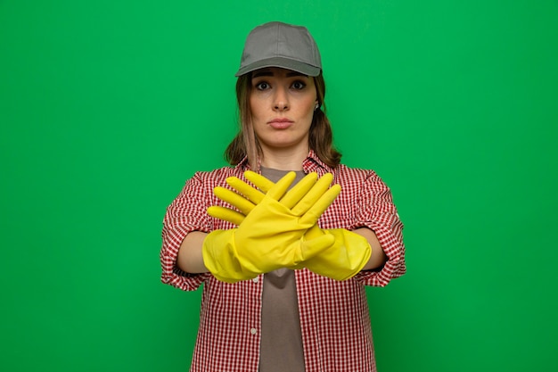 Jonge schoonmaakster in geruit hemd en pet met rubberen handschoenen die naar de camera kijkt met een serieus gezicht dat een stopgebaar maakt met handen die over een groene achtergrond staan