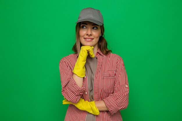 Jonge schoonmaakster in geruit hemd en pet met rubberen handschoenen die opzij kijkt met een glimlach op het gezicht met een peinzende uitdrukking