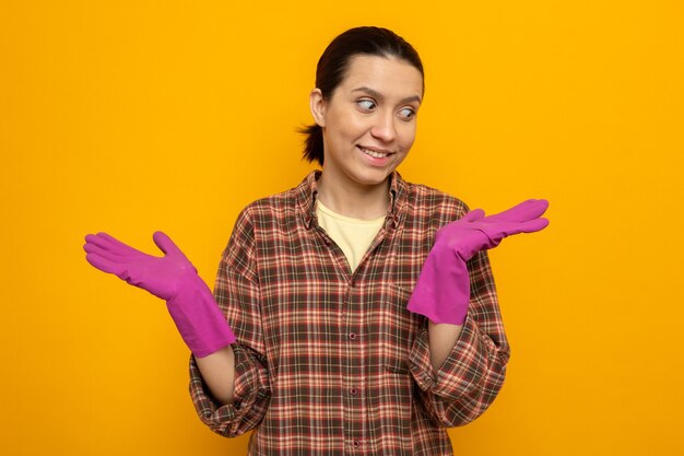 Jonge schoonmaakster in geruit hemd in rubberen handschoenen die opzij kijkt, blij en positief opvoedende armen vrolijk glimlachen