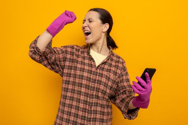 Jonge schoonmaakster in geruit hemd in rubberen handschoenen die smartphone blij en opgewonden houdt en vuist op oranje houdt