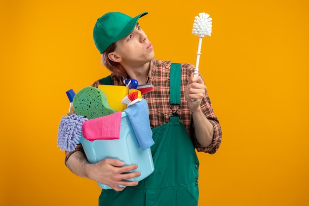 Jonge schoonmaakster in geruit hemd jumpsuit en pet met schoonmaakborstel en emmer met schoonmaakgereedschap kijkend naar borstel geïntrigeerd staande over oranje muur