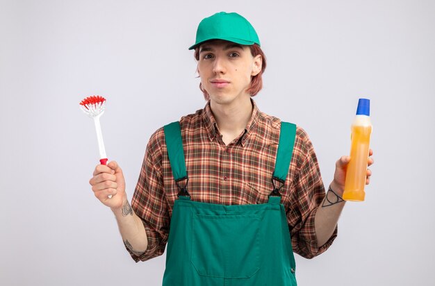 Jonge schoonmaakster in geruit hemd jumpsuit en pet met schoonmaakborstel en fles met schoonmaakspullen die er zelfverzekerd glimlachend uitziet