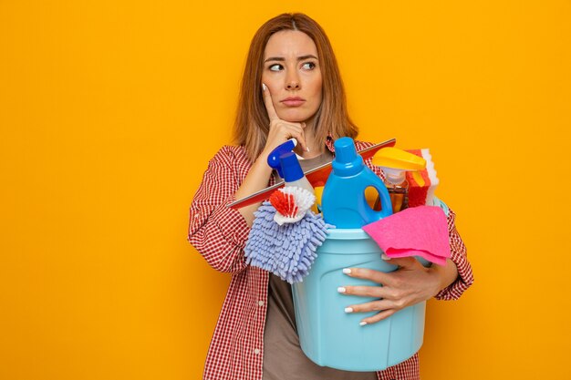 Jonge schoonmaakster in geruit hemd met emmer met schoonmaakhulpmiddelen die verbaasd opzij kijken