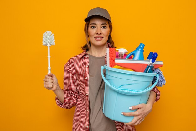 Jonge schoonmaakster in plaidoverhemd en GLB die emmer met schoonmakende hulpmiddelen en schoonmaakborstel houden die vrolijk gelukkig en positief klaar glimlachen