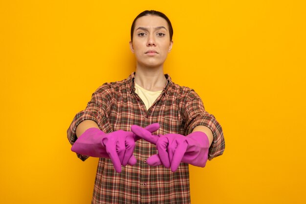 Jonge schoonmaakster in vrijetijdskleding in rubberen handschoenen die met een serieus gezicht kijkt en een verdedigingsgebaar maakt terwijl ze de wijsvinger kruist