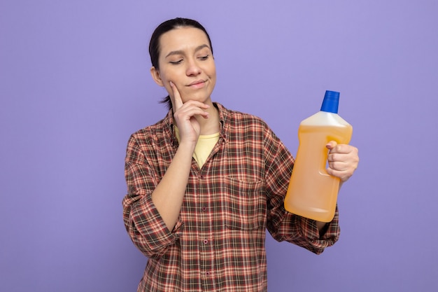 Jonge schoonmaakster in vrijetijdskleding met een fles schoonmaakspullen die ernaar kijkt met een peinzende uitdrukking op een paarse achtergrond