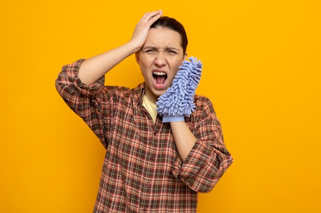 Jonge schoonmaakster in vrijetijdskleding met een stofdoek die schreeuwt met een geïrriteerde uitdrukking die gefrustreerd staat over de oranje muur