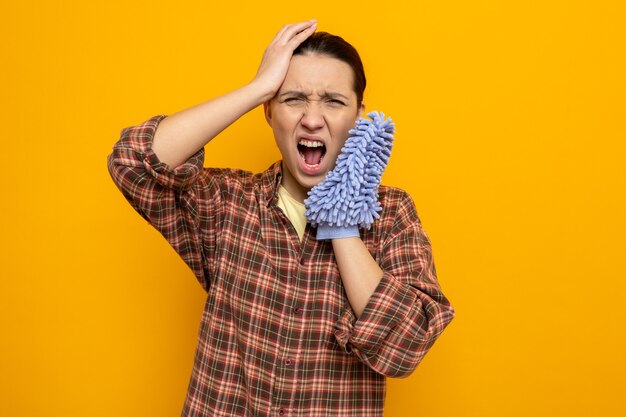 Jonge schoonmaakster in vrijetijdskleding met een stofdoek die schreeuwt met een geïrriteerde uitdrukking die gefrustreerd staat over de oranje muur