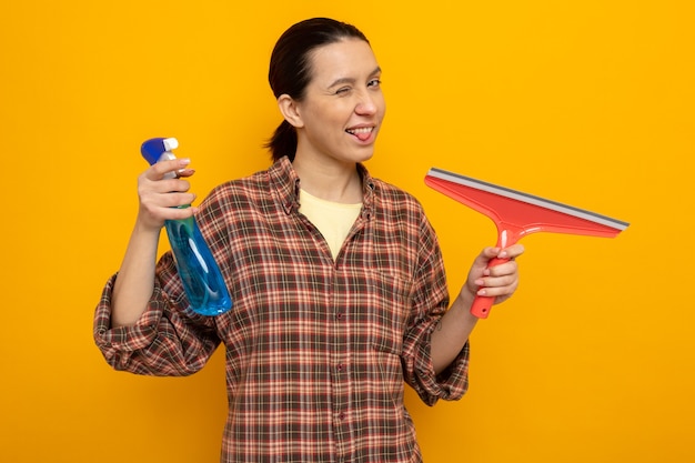 Jonge schoonmaakster in vrijetijdskleding met schoonmaakspray en dweil kijkend naar de voorkant, blij en positief, tong uitsteekt die over oranje muur staat