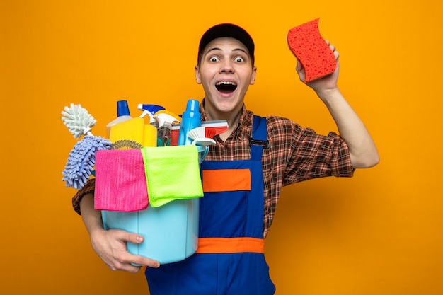 Jonge schoonmaakster met uniform en pet met emmer schoonmaakgereedschap met spons geïsoleerd op oranje muur