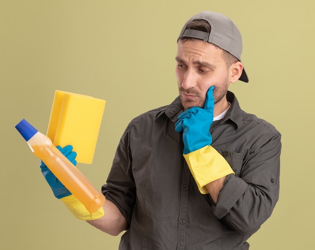 Jonge schoonmakende mens die vrijetijdskleding en GLB in rubberhandschoenen draagt die fles met schoonmakende levering en spons houdt die het met peinzende uitdrukking bekijkt die zich over groene muur bevindt