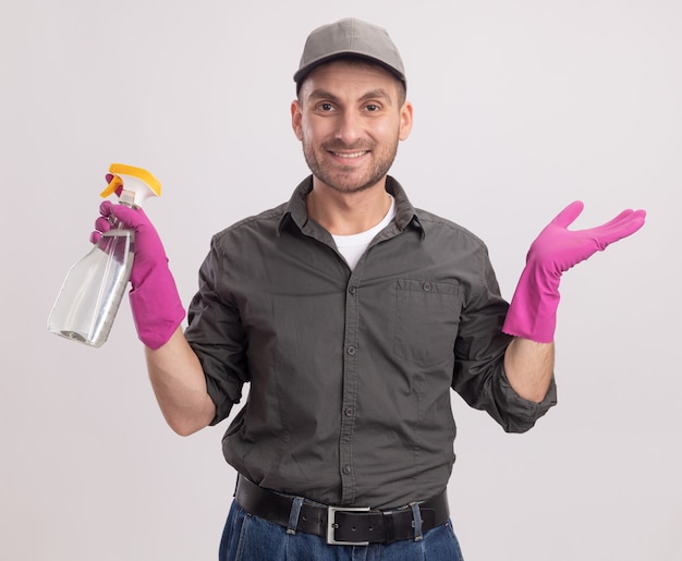 Jonge schoonmakende mens die vrijetijdskleding en GLB in rubberhandschoenen draagt die nevelfles houdt die vrolijk over witte muur glimlacht