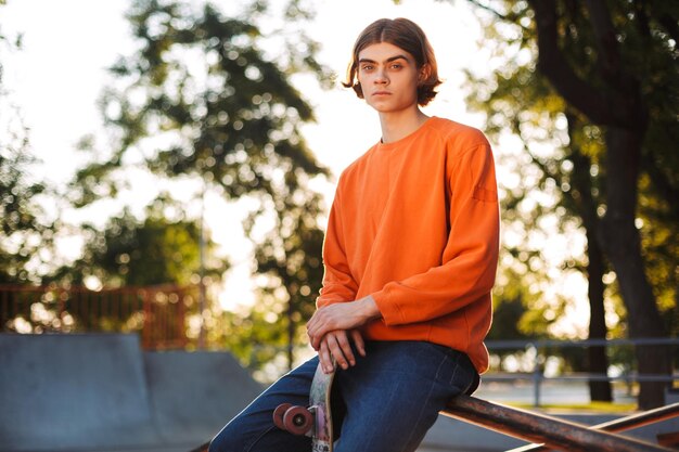 Jonge serieuze skater in oranje pullover die bedachtzaam in de camera kijkt met skateboard terwijl hij op de reling in het moderne skatepark leunt