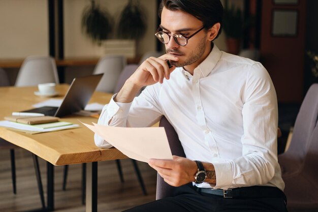 Jonge serieuze zakenman in een bril die zorgvuldig papieren leest terwijl hij in een modern kantoor werkt
