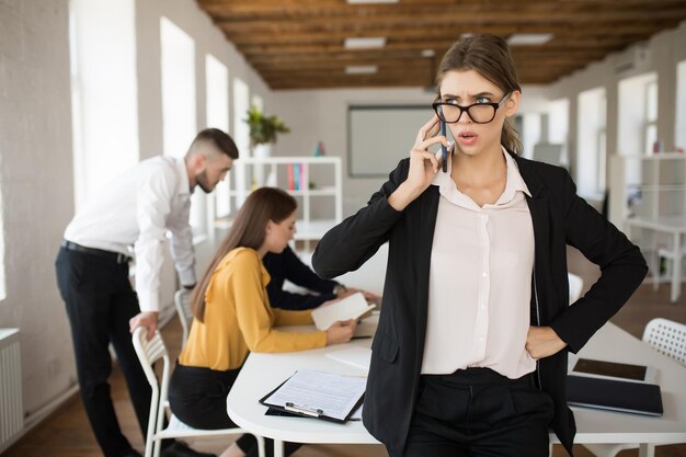 Jonge serieuze zakenvrouw in bril en shirt die verbaasd opzij kijkt terwijl ze op mobiel op kantoor praat met collega's op de achtergrond