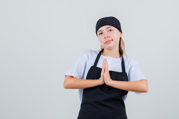 Jonge serveerster die namaste-gebaar in uniform en schort toont en geestloos kijkt