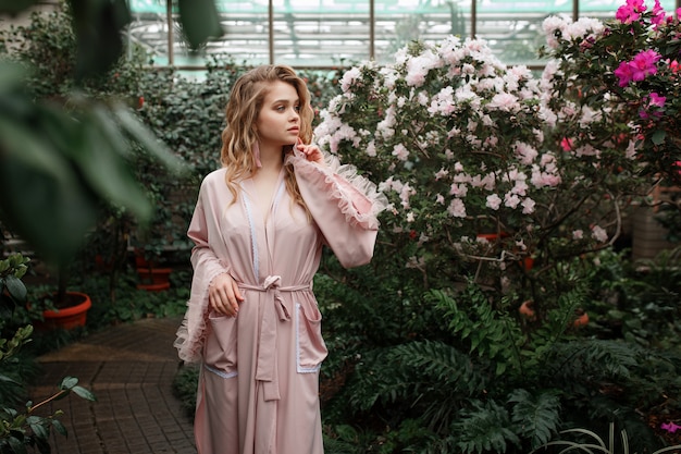 jonge sexy vrouw in roze badjas staande in de ochtend tuin vol met bloemen