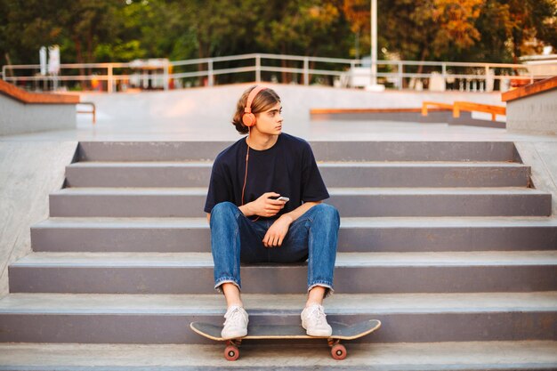 Jonge skater in oranje koptelefoon met mobiele telefoon in handen zittend op trappen in modern skatepark