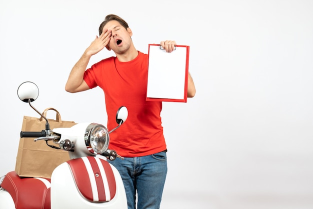 Jonge slaperige bezorger in rode uniform staande in de buurt van scooter met document op witte muur