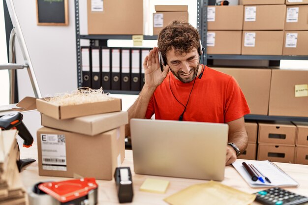 Jonge spaanse callcenter agent man aan het werk in het magazijn glimlachend met hand over oor luisterend naar geruchten of roddels. doofheid concept.
