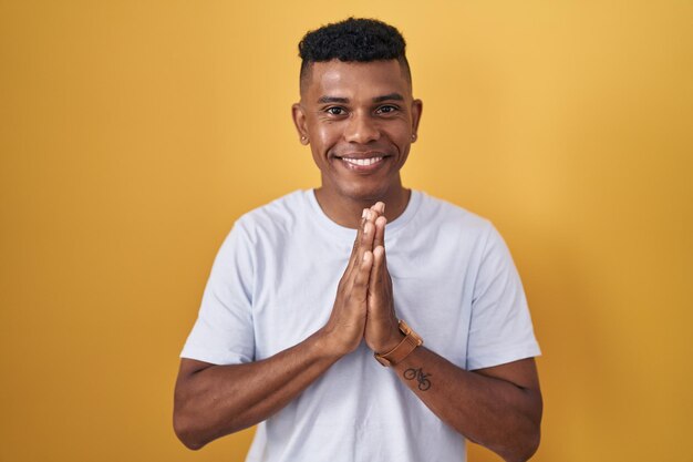 Jonge spaanse man die over een gele achtergrond staat en bidt met de handen samen om vergeving te vragen en zelfverzekerd glimlacht