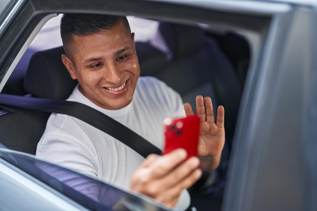 Jonge spaanse man doet videogesprek met smartphone in de auto die er positief en gelukkig uitziet en glimlacht met een zelfverzekerde glimlach die tanden laat zien