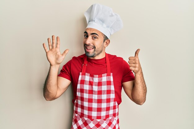 Jonge spaanse man in bakkersuniform die toont en omhoog wijst met vingers nummer zes terwijl hij zelfverzekerd en gelukkig glimlacht