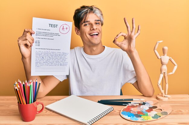 Jonge spaanse man kunstenaar die geslaagd is voor examen zittend op de tafel doet ok teken met vingers glimlachend vriendelijk gebarend uitstekend symbool